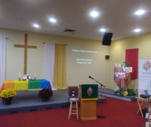 Sanctuary with a wooden cross on the wall, a rainbow cloth on the communion table, a stool, and a pulpit with the United Church crest on it. 