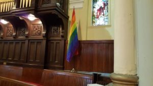 A rainbow flag stands between a stained glass window and organ pipes with intricately carved wood, in a church worship space. 
