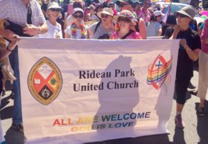 A group of smiling people stand in the sun behind a white banner with the United Church logo on the left and Affirm's logo on the right. It reads: "Rideau Park United Church, all are welcome. God is love". 