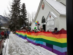A huge rainbow banner stretches along the front of a white clapboard church, held up by dozens of people. 